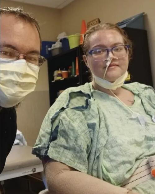 A man in a face mask beside a woman in a hospital gown with an oxygen tube and bandages on her arms.