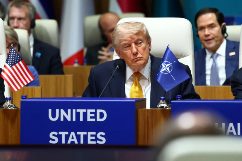 Donald Trump at a conference table with a NATO flag and a "UNITED STATES" placard in front of him.