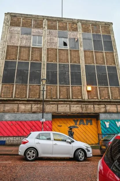 A boarded-up and derelict building in Dudley with a parked white car and a pedestrian with an umbrella.