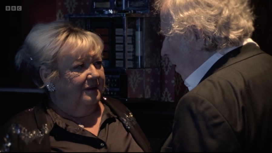 An older woman with blonde hair, wearing a sequined top and silver earrings, looking at a man with curly blonde hair.