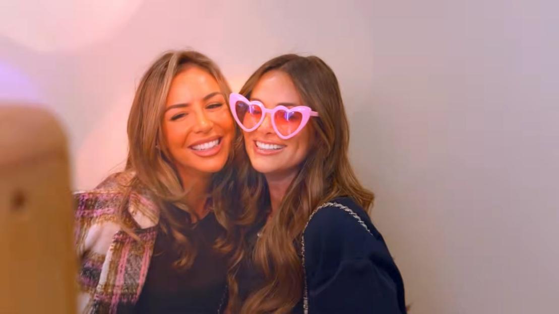 Two smiling women posing for a photo, one wearing pink heart-shaped glasses.