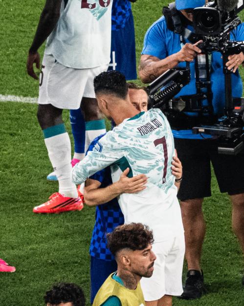 Football player Cristiano Ronaldo in a light blue and white jersey with "RONALDO 7" on the back, hugging a player in a blue checkered uniform on a green field.