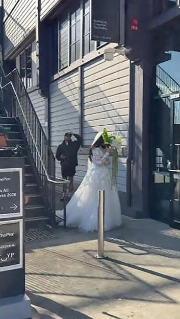 A bride in a white gown and veil holds a bouquet, standing outside a building as a person watches from a staircase.