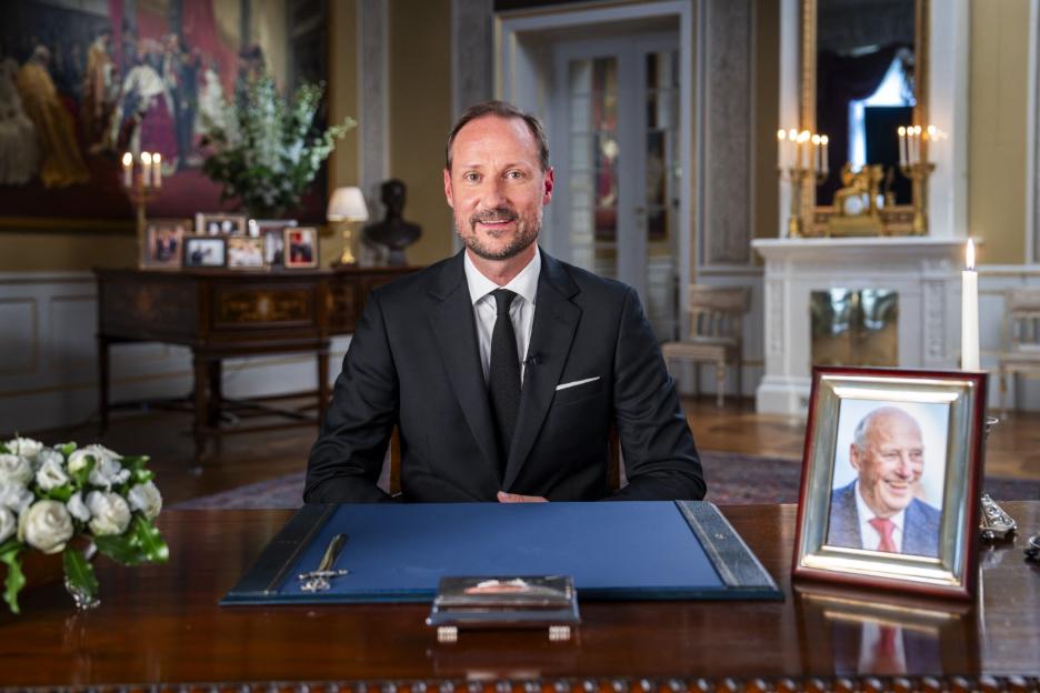 Crown Prince Haakon of Norway sitting at a desk in a formal room, with a framed photo of King Harald V to his right.
