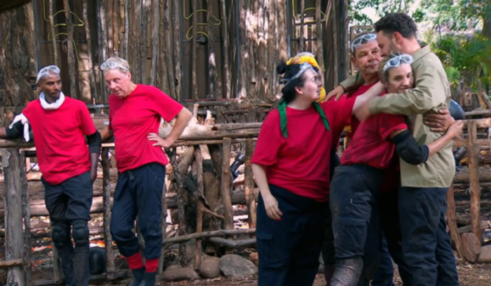 People in red shirts in a jungle camp, with some embracing and others standing.