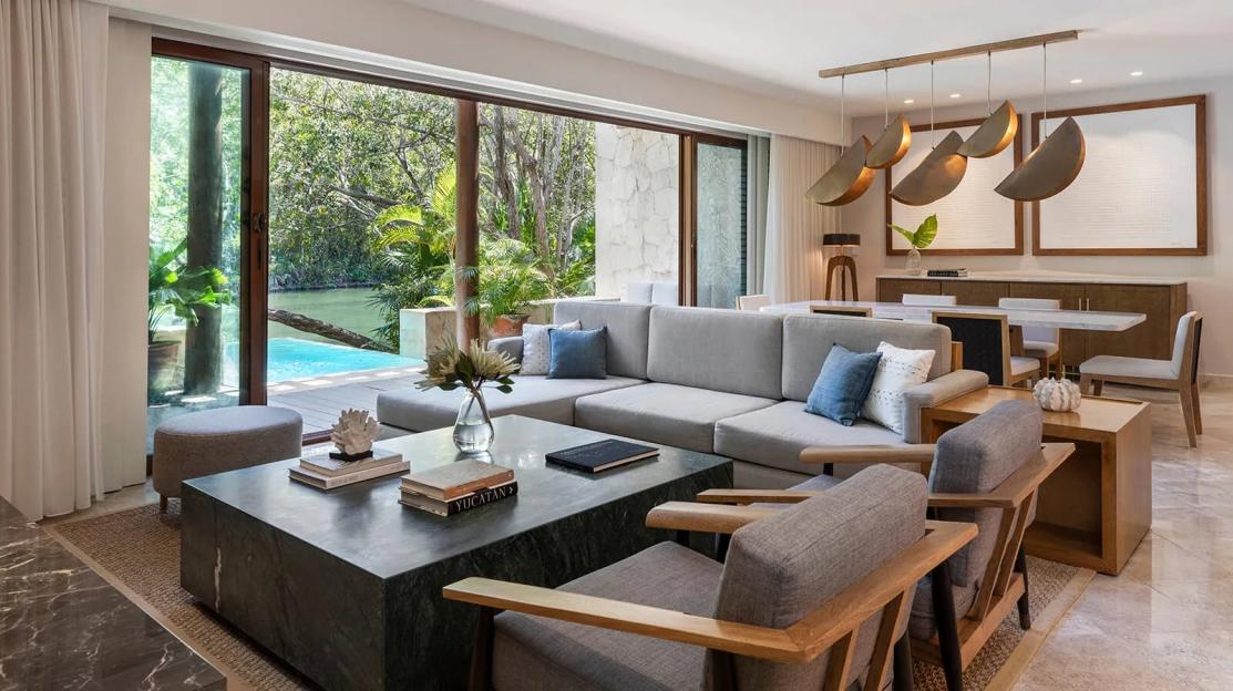 A modern living room with a gray sectional sofa, chairs, a large stone coffee table, and a dining area, featuring a large glass door overlooking a pool and lush greenery.