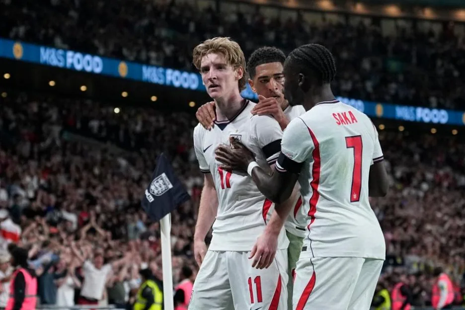 Three England football players in white jerseys with red numbers celebrate on the field, with "M&S FOOD" visible on an advertising board and fans in the background.