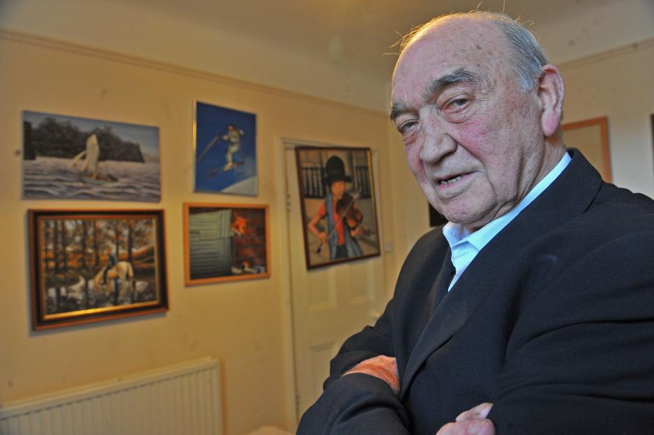 Eddie Richardson, a 60s gang boss, in a dark suit, stands in a room with five of his paintings on the wall.
