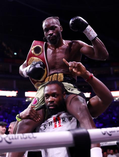 Dereck Chisora holding a championship belt while being carried on another man's shoulders.