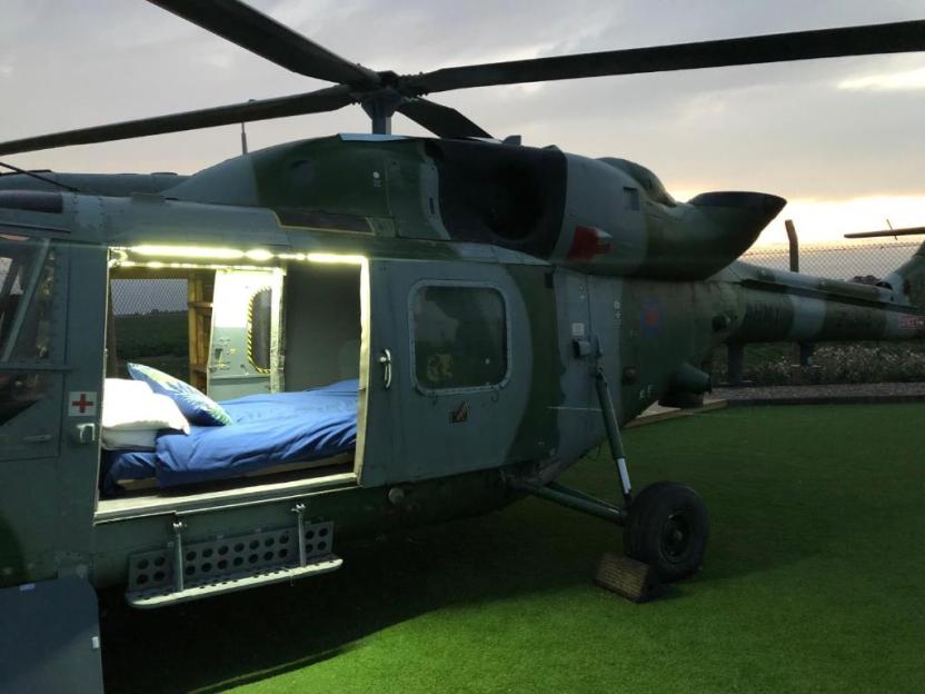 A helicopter converted into a sleeping accommodation with a bed and blue sheets visible inside, parked on green turf.