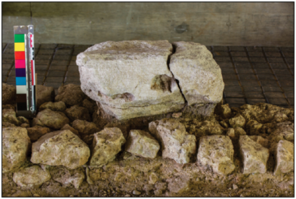 Archaeological stone remains with a crack, resting on a bed of smaller stones, next to a color calibration chart.