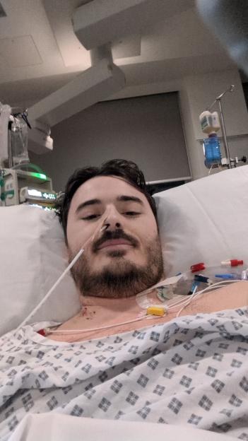 A man with a feeding tube in his nose and medical tubes coming from his chest lies in a hospital bed.
