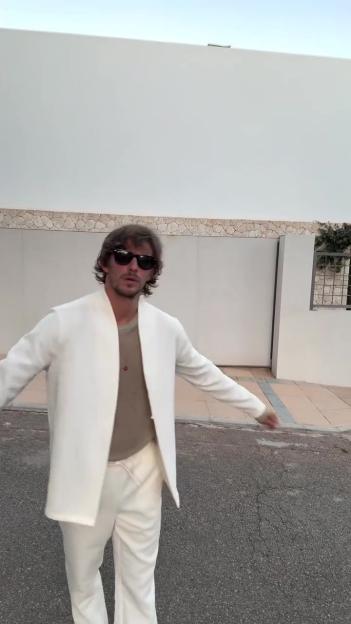 Man wearing sunglasses, a white jacket, and white pants, standing on asphalt.