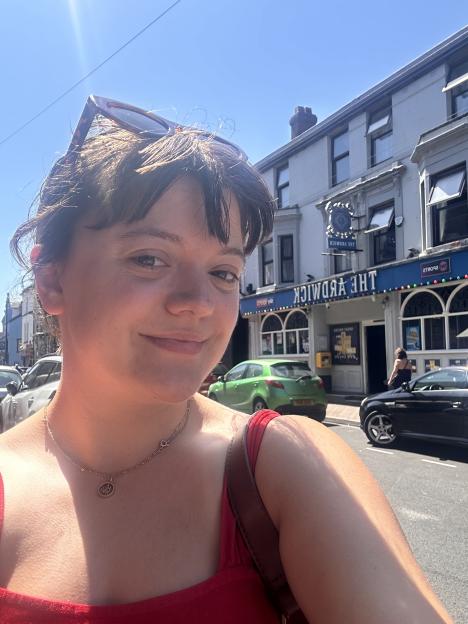A woman in a red top smiles while posing for a selfie in front of the Ardwick Pub.