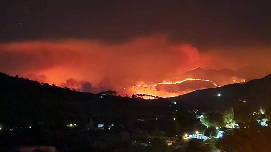 Nighttime wildfire blazing across a dark mountain range, casting an orange glow on the sky.