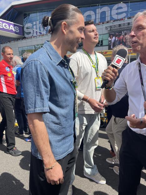An interviewer speaking into a microphone to a man wearing a denim shirt, with another man in a white shirt standing behind him.