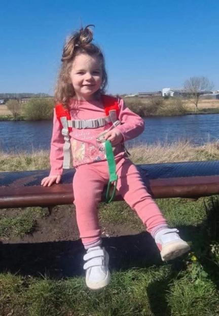 Isabelle Rose Welsh, a young girl with curly hair and a pink outfit, sitting on a bench with a body of water behind her.