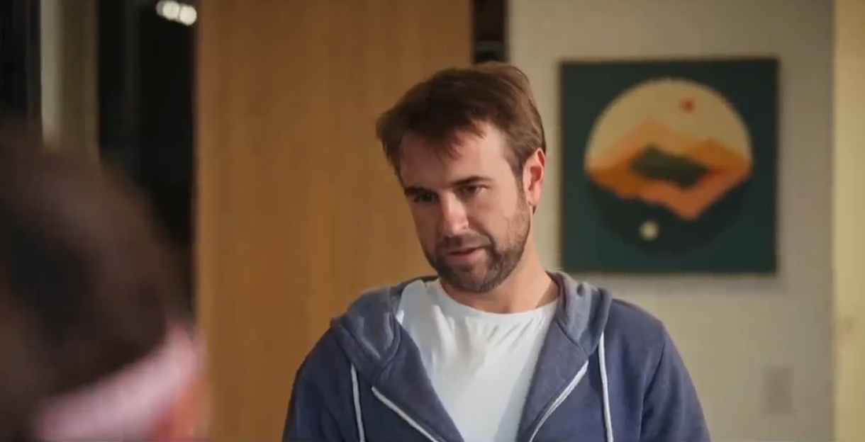 A man with a beard, wearing a white shirt and blue hoodie, looks to his left.