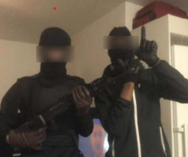 Two masked individuals, one holding an assault rifle and the other making a hand gesture.