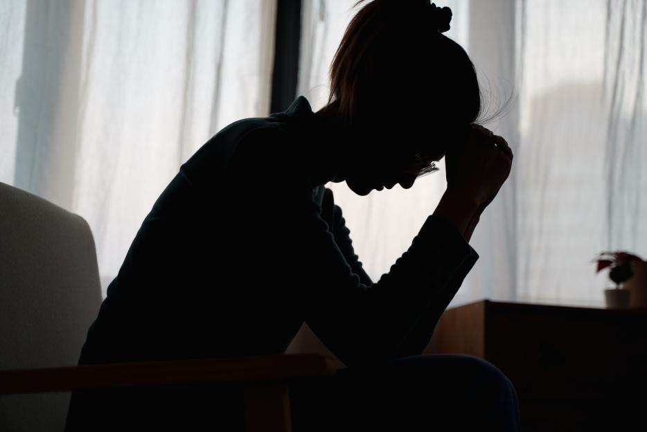 Silhouette of a young Asian woman sitting alone, head in hands, appearing stressed or sad, with a bright window in the background.