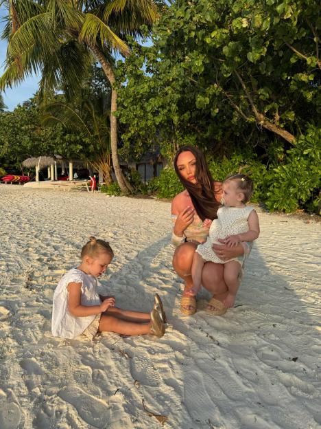 Charlotte Crosby holding a baby, with an older child playing on the sand beside them.