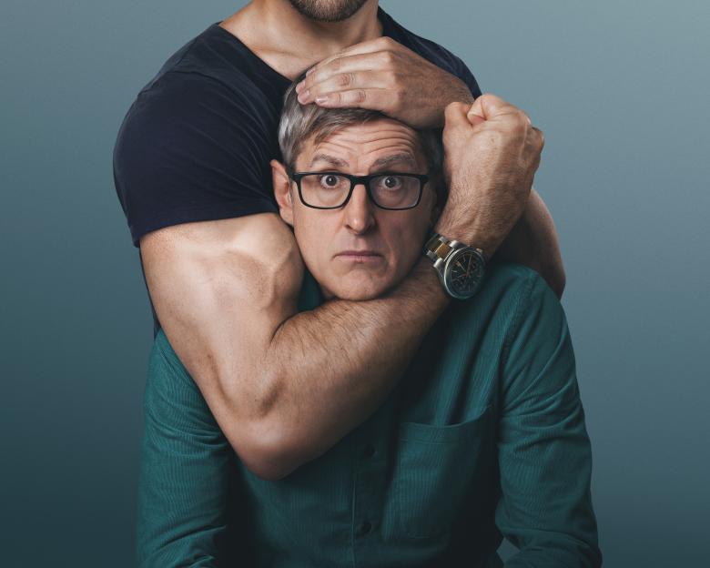 Louis Theroux's head held by the muscular arms of another person.