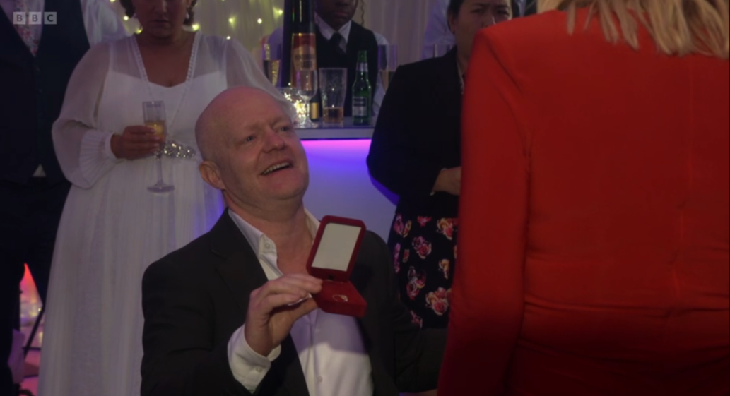 A man on one knee holding an open ring box to a woman in a red outfit, with other people in the background.