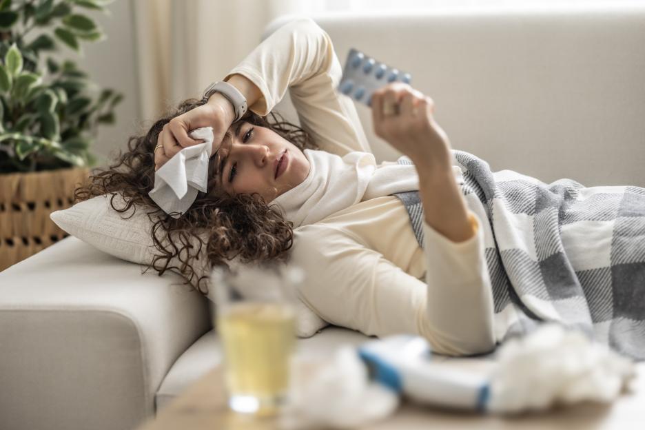 Sick young woman lying on the couch, holding a tissue to her forehead and a blister pack of pills.