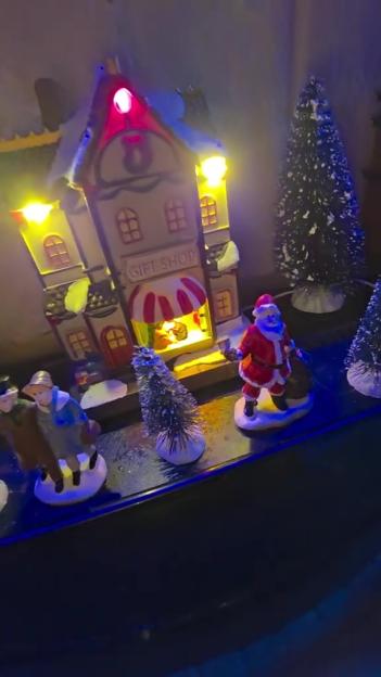 Illuminated Christmas village decorations with a gift shop, pine trees, and a Santa figurine.