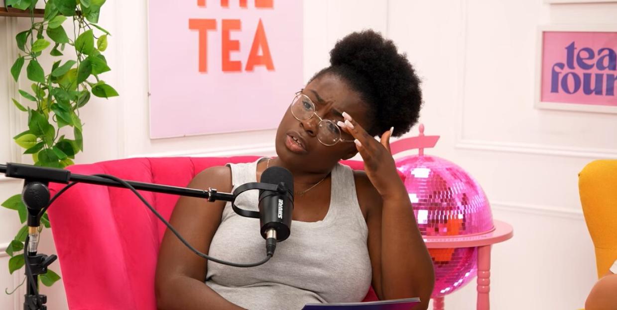 A Black woman with glasses in a grey tank top rubs her eye in a hot pink armchair while a microphone is in front of her.