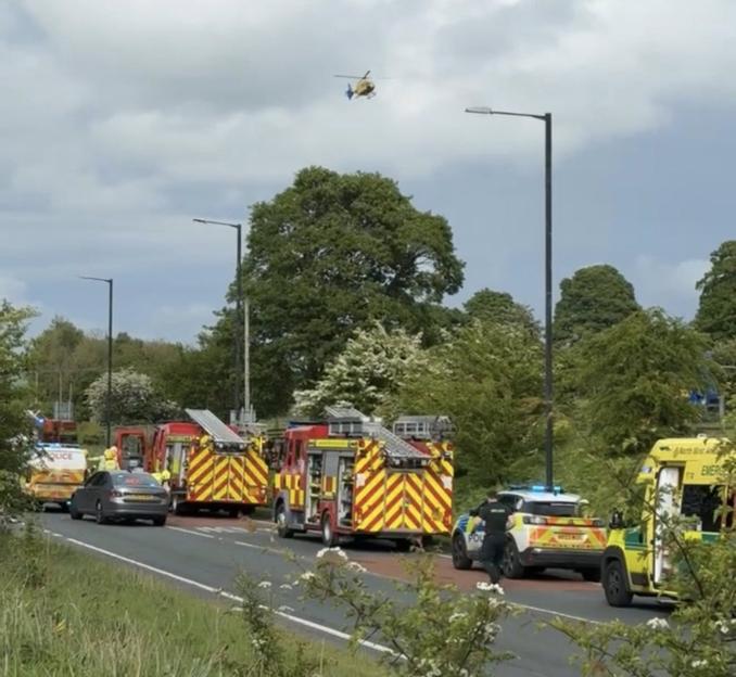 Emergency vehicles, including a police car, fire trucks, and ambulances, on a road with a helicopter flying overhead.