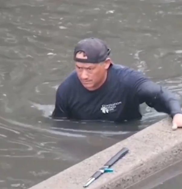 A man standing in floodwaters, looking towards an object on a concrete ledge.