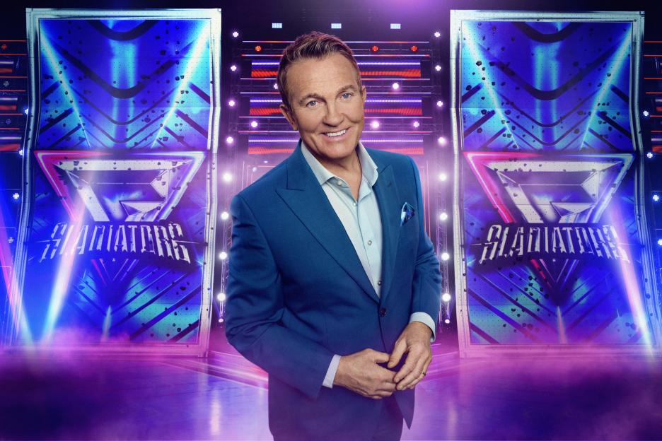 Bradley Walsh in a blue suit smiles in front of two large Gladiators logos.