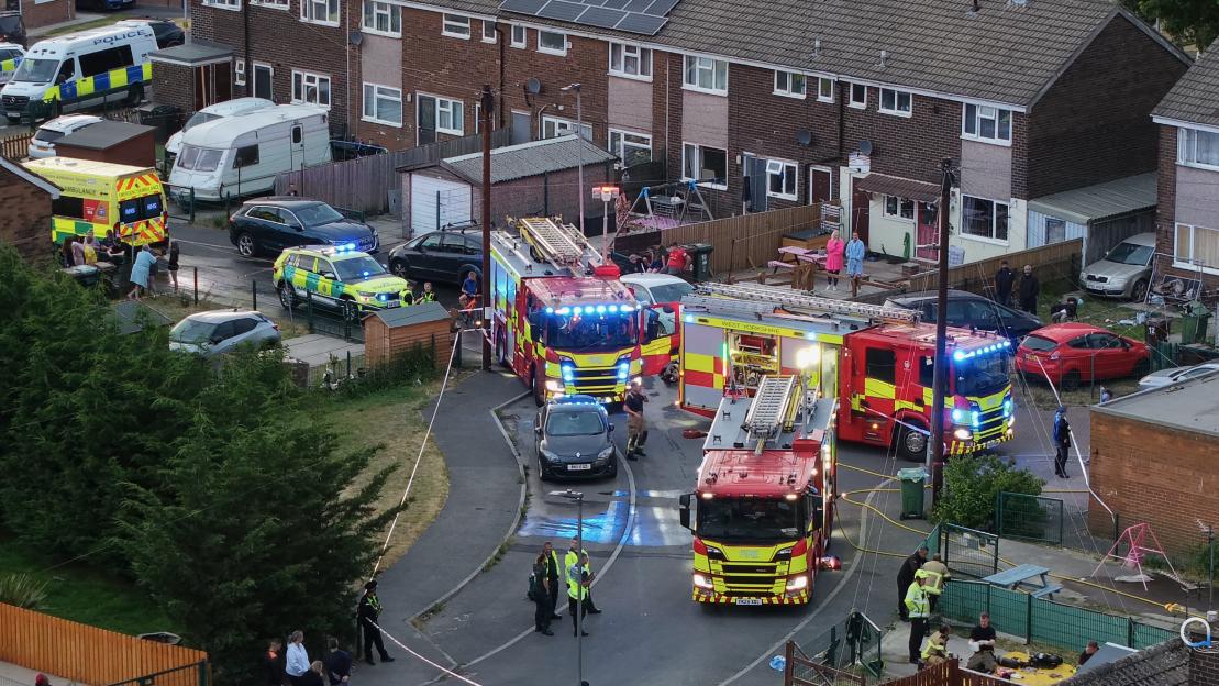 Emergency services respond to a house fire, with fire trucks, an ambulance, and police vehicles present.