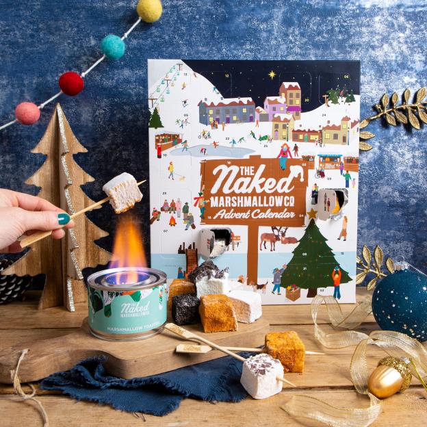 A hand holding a toasted marshmallow on a stick over a Naked Marshmallow Co. toaster, next to a Naked Marshmallow Co. Advent Calendar filled with different flavored marshmallows.
