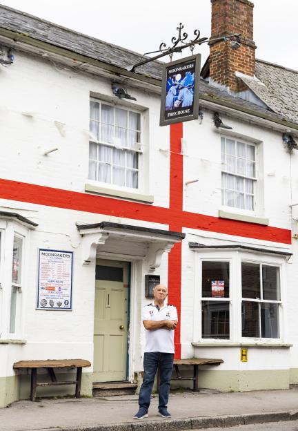 A man standing in front of a white pub with a large red St. George's Cross painted on its exterior, and the pub sign "Moonrakers Free House".