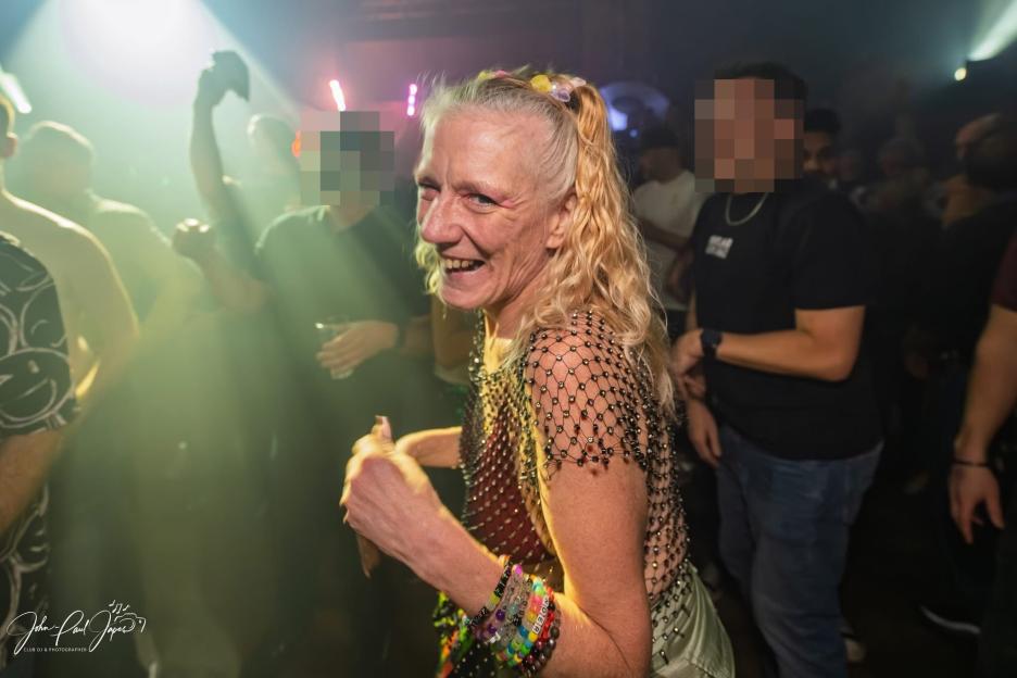 A smiling woman with blonde hair, wearing a mesh top and beaded bracelets, dances at a rave.