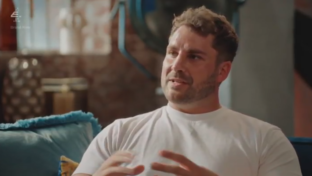 A man with short brown hair and a beard, wearing a white t-shirt, sits on a blue couch and gestures with his hands while speaking.