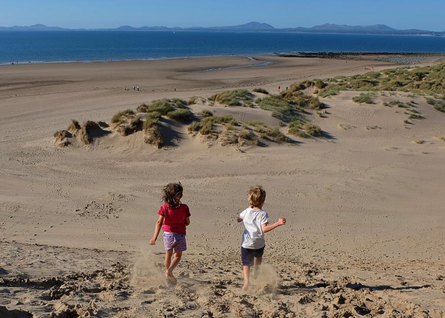Two children running down a sand dune towards the beach and ocean at Shell Island, Wales.