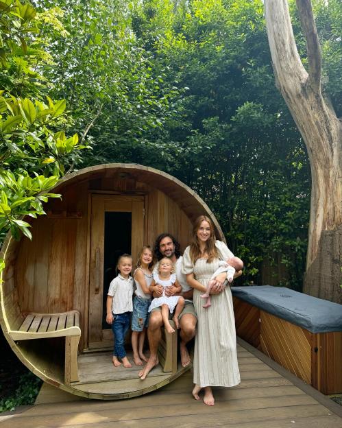 A family of six, including a newborn, posing barefoot in front of a wooden sauna and hot tub.