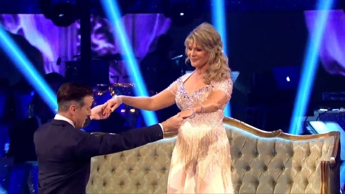 Ruth Langsford dancing with a partner on a stage with a blue velvet couch in the foreground.