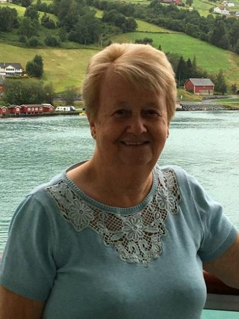 June Henty smiling in front of a body of water with buildings and green hills in the background.