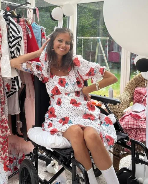 Deborah James smiles while sitting in a wheelchair, wearing a white dress with red poppies.