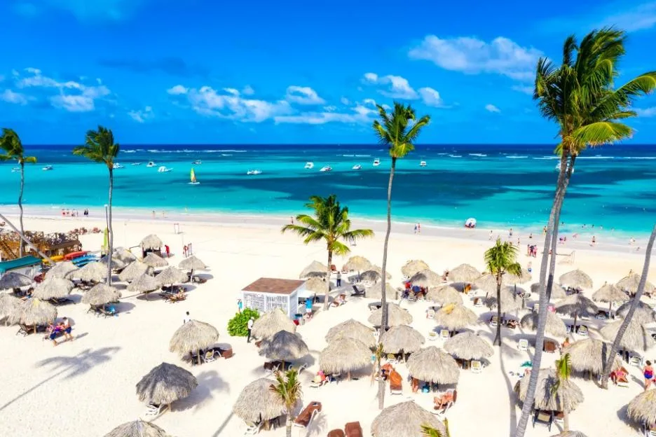 Aerial view of Bavaro Beach in Punta Cana with white sand, palm trees, thatched umbrellas, and turquoise ocean water.
