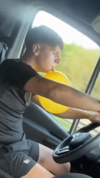 A young man inhales nitrous oxide from a yellow balloon while driving a van.