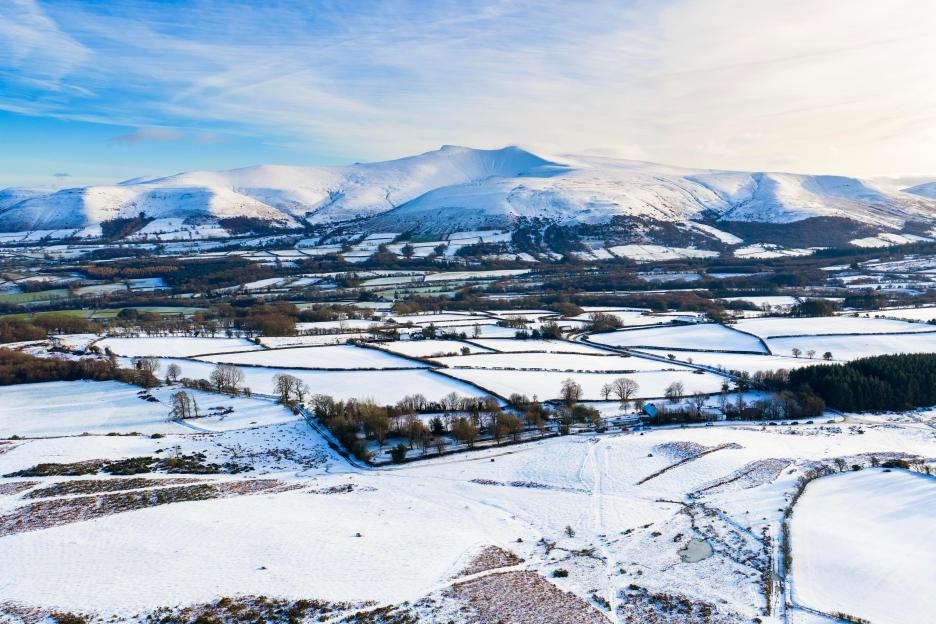 Aerial view of the snow-covered mountains of Bannau Brycheiniog National Park, with fields and trees in the foreground.