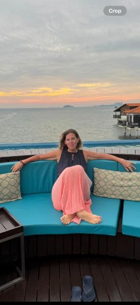 Shona Sibary sitting on a blue outdoor sofa, wearing a dark blue top and a light pink skirt, with the ocean and sunset in the background.