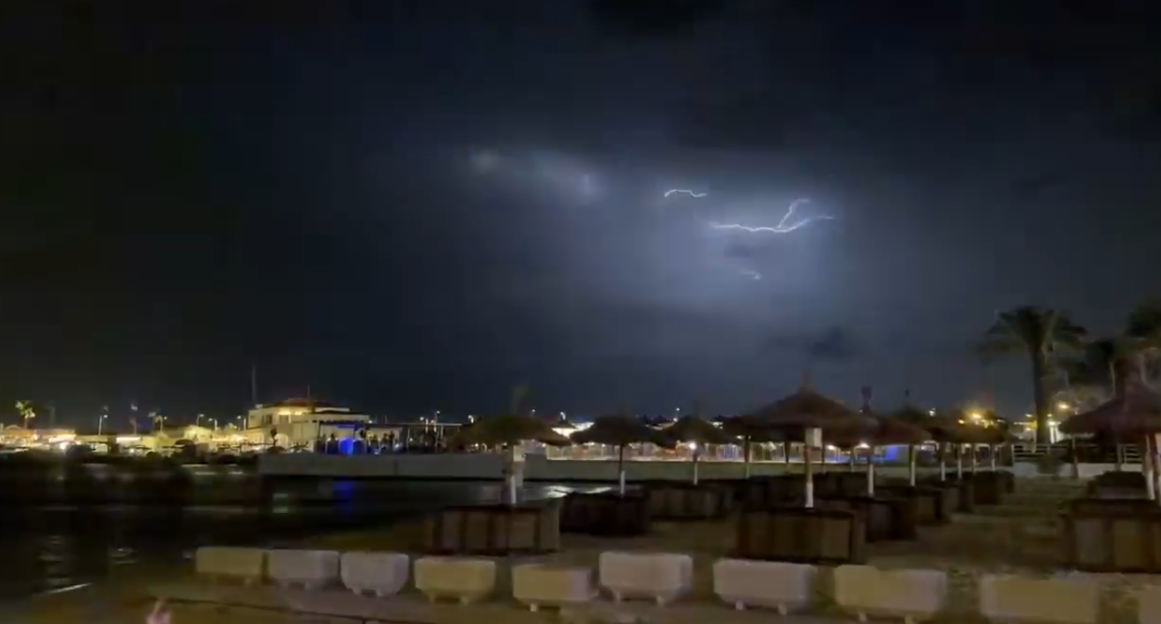 Lightning flashes across a dark sky over a beach with straw umbrellas and distant buildings at night.
