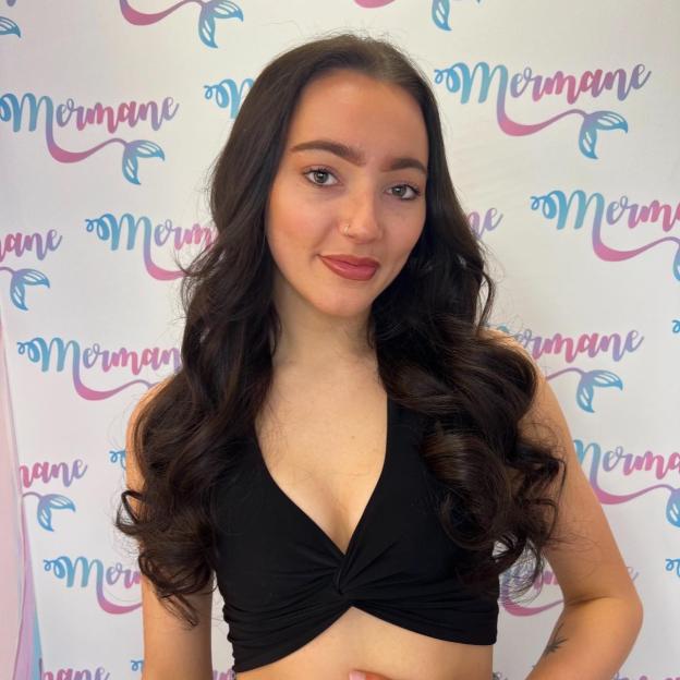 Jess Waterman, who died in a car crash at 20, shown smiling with long dark hair, wearing a black top, against a white background with a "Mermane" logo.