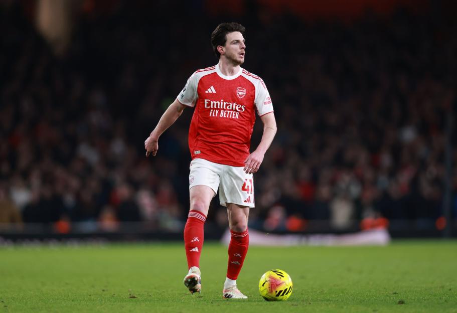 An Arsenal player in a red and white uniform standing on a soccer field with a ball next to his foot.